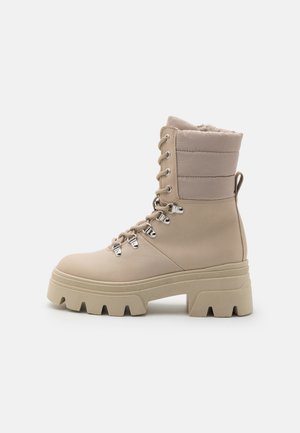 Botas beige con cordones y suela gruesa, sección superior acolchada, herrajes plateados y material texturizado. Presenta un diseño robusto y una construcción sólida.