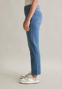 Hoge blauwe denim jeans met een rechte snit, voorzien van achterzakken, gedragen met beige en witte sneakers, tegen een lichte achtergrond.