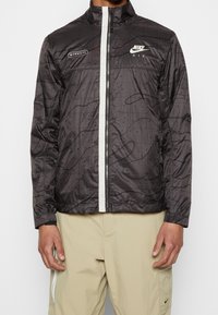 Musta Nike Air -takki, jossa on vetoketjukiinnitys, korkea kaulus, hienovarainen kohokuvioitu kuvio ja valkoiset yksityiskohdat vetoketjussa ja brändäyksessä.