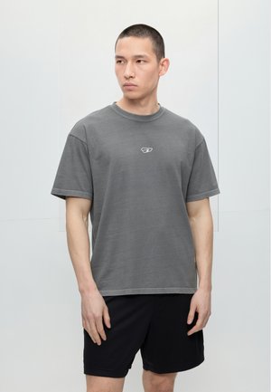 Άνδρας με κοντά μαλλιά που φοράει γκρι φαρδιά μπλούζα με λογότυπο και μαύρα σορτς, στέκεται μπροστά σε μια απλή φωτεινή τοίχο κοιτάζοντας πλευρικά.