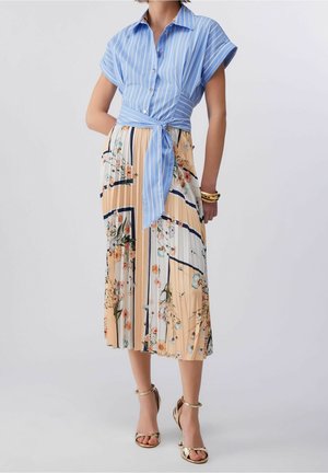 Vrouw draagt een blauw gestreept kort mouw blouseje, geknoopt in de taille, en een geplooide bloemen midi rok met gouden hoge hak sandalen.