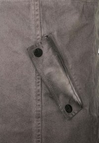 Grauer Stoff mit glatter Textur, versehen mit einer seitlichen Tasche, die mit einem schwarzen Knopf gesichert ist. Sichtbare Nähte entlang der Kanten.