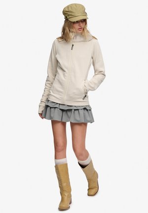 Ung kvinde iført beige lynlåstrøje, grå flæset nederdel, tanfarvede støvler, hvide sokker og olivengrøn kasket stående med den ene hånd i lommen.