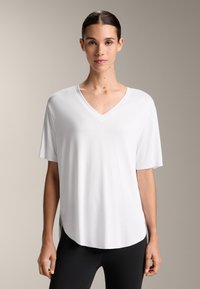 Bílý tílko s V výstřihem a krátkými rukávy, vyrobené z hladkého materiálu. Volný střih s zaobleným lemem, v kombinaci s černými legínami. Jednoduché a všestranné.