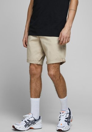 Mann trägt beige hochgekrempelte Shorts, schwarzes T-Shirt, weiße Kniestrümpfe und weiße Laufschuhe mit marineblauen und orangen Akzenten, steht auf grauem Boden.