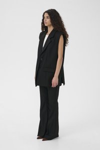 Must musta-ülikonnajakk struktureeritud disainiga, ühe rea nööpidega ja varrukateta, koos mustade laienevate pükste ning teravate pruuni värvi kingadega.