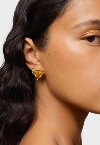 Orecchino a perno in tonalità oro con una pietra gialla a forma triangolare, caratterizzato da una superficie liscia e una finitura lucida, indossato all'orecchio.
