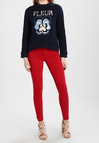 Felpa navy con testo "FLEUR" e design floreale, abbinata a jeans rossi aderenti e tacchi nudi con fascette. Tessuto liscio, design casual.