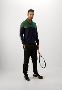 Costum sport verde și bleumarin, jachetă cu fermoar și dungi laterale, pantaloni negri cu logo, asortați cu adidași albaștri și albi, ținând o rachetă de tenis.