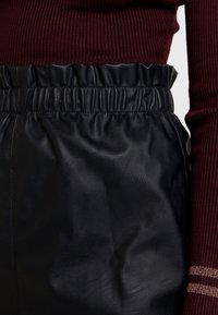 Falda de cuero negro con una cinturilla elástica, con una textura suave. Parte superior acanalada en color burdeos parcialmente visible con detalles dorados.