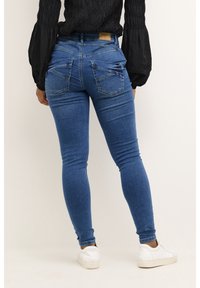 Jeans ajustados de mezclilla azul con una textura suave, que cuentan con bolsillos traseros y una cintura alta, combinados con zapatillas blancas y una camiseta negra.