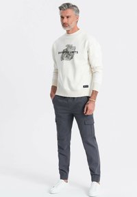 Cremefarvet sweatshirt med grafisk tryk og teksten "SMASHING LIMITS", parret med mørkegrå cargo bukser og hvide sneakers.
