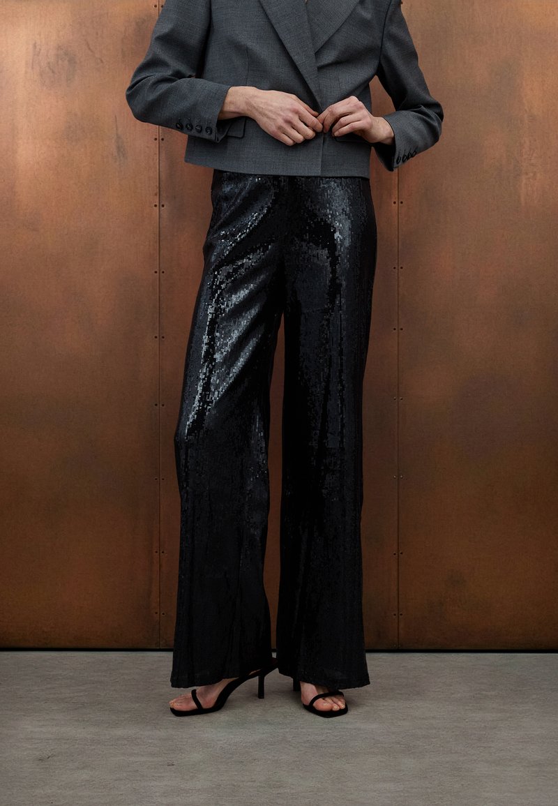 Μαύροι φαρδιοί παντελόνες με παγιέτες και γυαλιστερό φινίρισμα, σε συνδυασμό με cropped γκρι σακάκι. Το μοντέλο φοράει μαύρα σανδάλια. Το φόντο είναι ένας υφασμάτινος χάλκινος τοίχος.