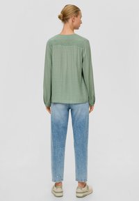 Lichtgroen langemouwen shirt met getextureerde details op de schouders, gecombineerd met hoge taille blauwe jeans en beige sneakers.