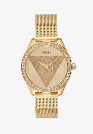Montre en métal doré avec un bracelet en maille, un cadran triangulaire en texture sable et une lunette décorée de pierres. Elle présente des aiguilles dorées et le logo 'Guess'.