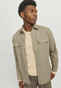 Chemise boutonnée gris clair avec deux poches poitrine, portée sur un pull beige. Coupe droite, tissu doux et détails de texture subtils.