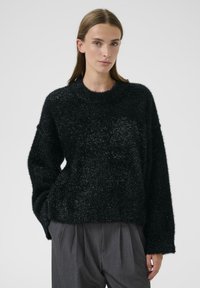 Czarny, puszysty sweter o okrągłym dekolcie i długich rękawach, charakteryzujący się fakturowaną powierzchnią i luźnym krojem, w połączeniu z szarymi spodniami plisowanymi.