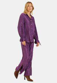 Fioletowy satynowy zestaw piżamowy z teksturowanym wzorem w diamenty, długie rękawy z akcentami w postaci kokardek oraz bocznymi rozcięciami w spodniach. Brązowe obcasy uzupełniają outfit.