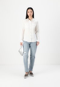 White button-up shirt with two chest pockets, light-wash ripped jeans, silver metallic clutch, and silver embellished shoes.