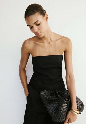 Mujer con un top negro sin tirantes y pantalones, sosteniendo un gran bolso negro tejido, lleva pulseras doradas y mira hacia abajo.
