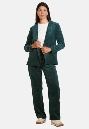 Completo in corda verde acqua composto da un blazer aderente con rever a forma di nota e pantaloni a gamba larga, abbinato a una t-shirt bianca e scarpe beidge.