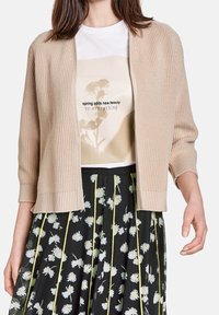 Béžový pletený svetr s tříčtvrtečními rukávy, nošený přes bílé tričko s květinovou grafikou, doplněný černou sukní s květinovým potiskem.