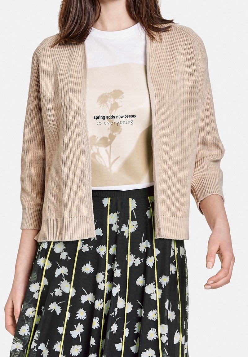 Béžový pletený svetr s tříčtvrtečními rukávy, nošený přes bílé tričko s květinovou grafikou, doplněný černou sukní s květinovým potiskem.