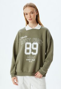 Sudadera verde oliva con cuello blanco y texto impreso. Cuello redondo, mangas largas, puños acanalados, con gráficos de "89" y "Florida WEST COAST".