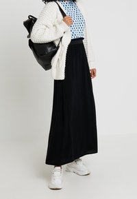 Gonna nera lunga con una texture morbida, abbinata a una blusa azzurro chiaro a pois e a un cardigan bianco chunky, completata da sneakers bianche e uno zaino nero.