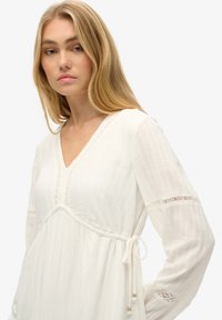 Vestido blanco de manga larga con escote en V, con tejido texturizado y detalles de encaje en las mangas. Cintura ajustable con cordón para un ajuste personalizado.