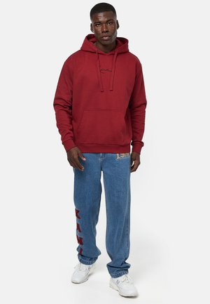 Bordeaux hættetrøje med sort logo på forsiden, parret med lysblå baggy denimjeans med rød tekst på venstre ben.