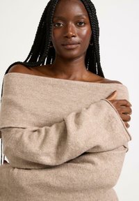 Beige off-shoulder tröja med vida ärmar, tillverkad av ett mjukt, texturerat tyg. Designen är lös och har en avslappnad fall.