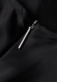 Czarny materiał o gładkiej fakturze wyróżnia się metalowym suwakiem w kolorze srebrnym, na którym widnieje napis "SUPERDRY," umiejscowionym w pobliżu dekoltu.
