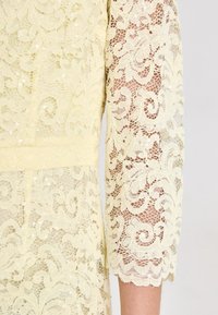 Robe en dentelle de couleur jaune pâle présentant des motifs floraux élaborés, des manches trois-quarts, et des accents en sequins sur un tissu texturé.