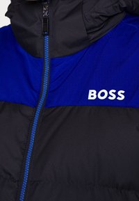 Puffová bunda v černé a modré látce, s zipovým uzávěrem, kapucí a výrazným bílým logem "BOSS" na hrudi. Strukturální materiál.