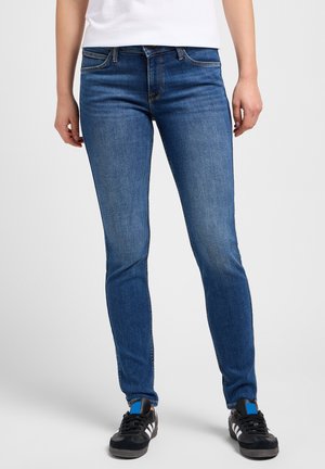 Person iført slim-fit blå jeans, sorte sneakers med hvide striber og en hvid indstukket top, stående mod en ensfarvet baggrund.