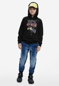 Zwarte hoodie met kleurrijk grafisch ontwerp, gecombineerd met vervaagde blauwe joggers met een band bij de enkel en zwarte sneakers. Gele pet gedragen.