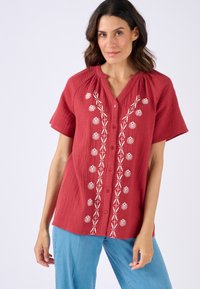 Camicia rossa con texture, maniche corte e scollo a V, caratterizzata da ricami floreali bianchi lungo la parte anteriore e diversi bottoni.