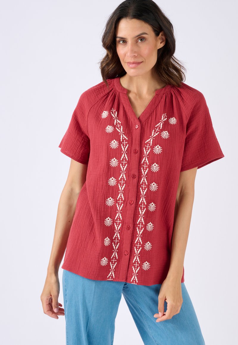 Camisa de tejido rojo con mangas cortas y escote en V, que presenta bordados florales blancos en la parte delantera y varios botones.