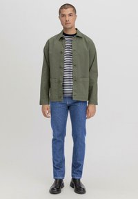 Olijfgroen katoenen jack met knopen, twee voorzakken, gedragen over een marineblauw en wit gestreept shirt, gecombineerd met blauwe jeans en zwarte laarzen.