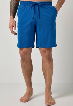 Hombre con pantalones cortos azules con cordón hasta la rodilla, de pie descalzo con las manos parcialmente visibles, mostrando tatuajes en las piernas y los brazos.