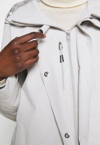 Světle šedá voděodolná bunda se zapínáním na zip, druky a hladkou texturou. Má kapuci a boční kapsy na zip.