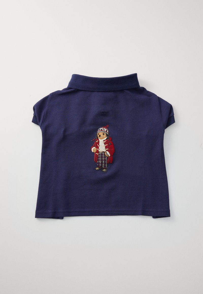 Marineblå polo-skjorte med korte ærmer, som har et brodert design av en figur i en rød frakk og mønstrete bukser foran.