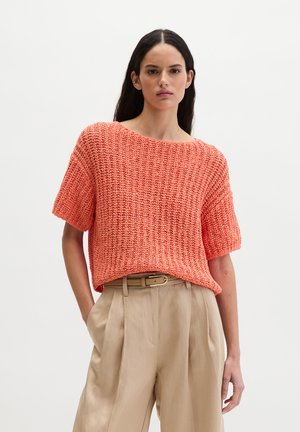 Donna con capelli lunghi e scuri che indossa un top corto di maglia corallo infilato in pantaloni beige a vita alta, plissettati con cintura.