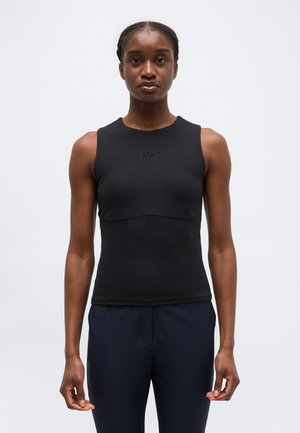 Γυναίκα που φοράει μαύρο αμάνικο ριμπ τοπ και σκούρο παντελόνι, στέκεται μπροστά σε απλό ανοιχτό γκρι φόντο, στραμμένη προς τα εμπρός.