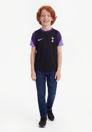 Garçon roux debout en souriant, portant un maillot de football noir et violet de Tottenham, un jean bleu et des baskets noires sur fond blanc.
