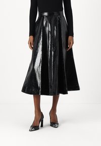 Falda midi plisada negra hecha de cuero sintético texturizado con acabado brillante que imita al cocodrilo, combinada con zapatos de tacón alto negros con acentos metálicos.