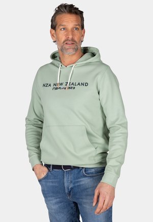 Groene hoodie met een vooraanstaande kangoeroezak en een trekkoordkap. Bevat de tekst "NZA NEW ZEALAND" en is gecombineerd met blauwe jeans.