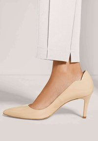 Beige Lackleder-Pumps mit spitzem Schuhdesign, kombiniert mit hellen, knöchellangen Hosen, die am Saum einen kleinen Schlitz haben.