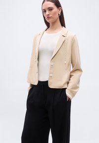 Γυναίκα με μπεζ κοντό σακάκι με λευκά κορδόνια, λευκή εφαρμοστή μπλούζα και μαύρο παντελόνι φαρδιάς γραμμής, στέκεται με τα χέρια στις τσέπες.